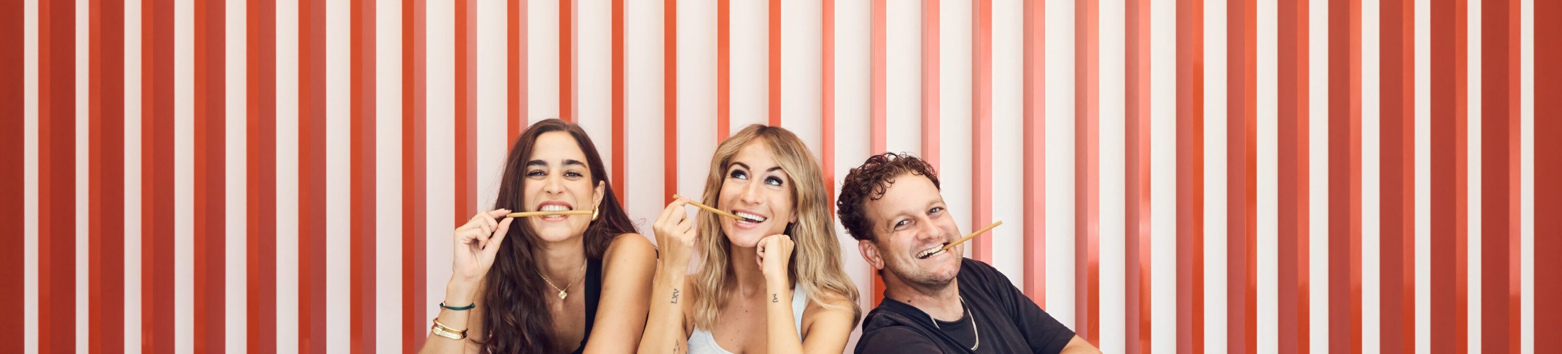 From left to right: Chiara Benz, Nadia Goedhart and Sandro Galfetti sitting side by side at a table, smiling, each playfully holding a golden Malbuner stick in their mouth. In the background are vertical red and white stripes.