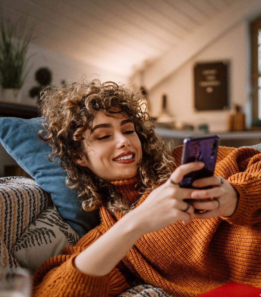 Frau liegt entspannt auf dem Sofa und schaut aufs Smartphone.